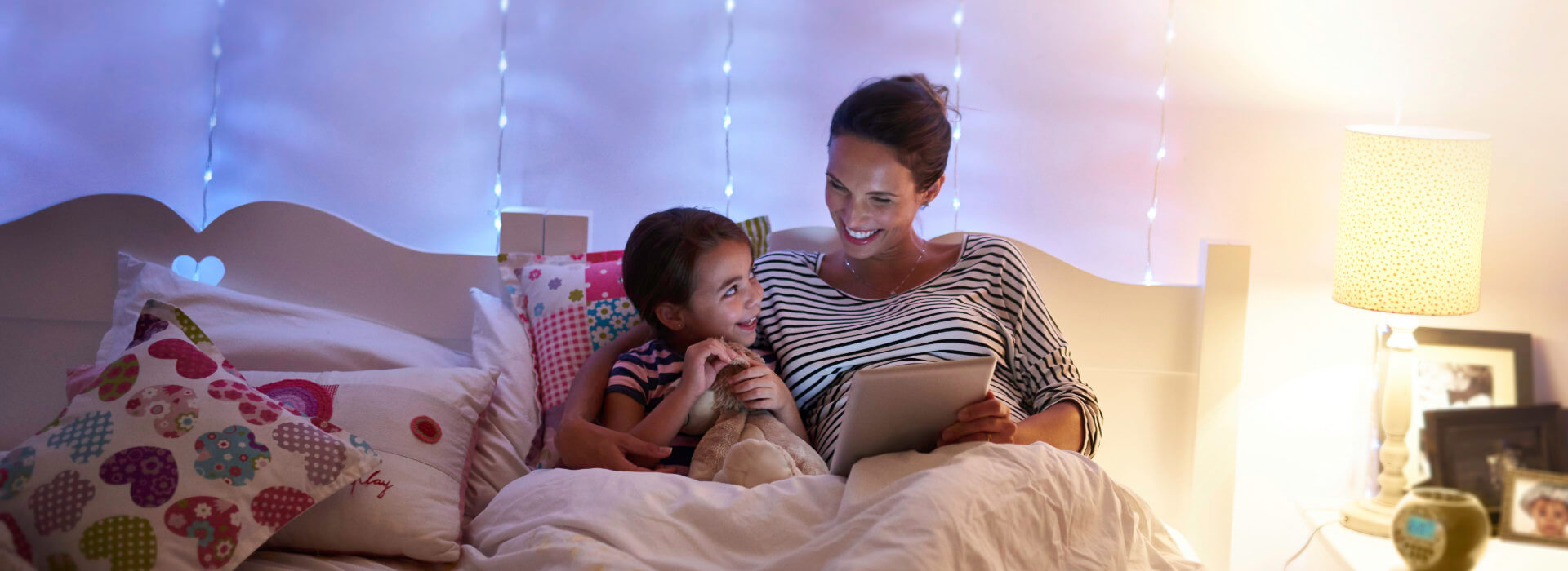 Mutter und Kind schauen im warm beleuchteten Kinderzimmer gemeinsam auf ein Tablet
