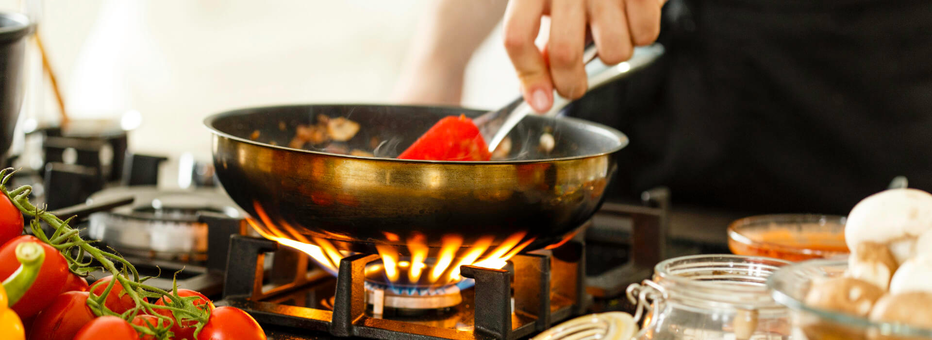 Pfanne auf einer Gasflamme, daneben Tomaten und Pilze